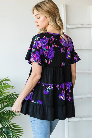 Woman wearing a black floral top with purple flowers, standing against a white wall.