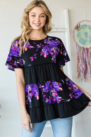 Woman wearing a black top with colorful floral patterns, standing in a room with a dreamcatcher in the background.