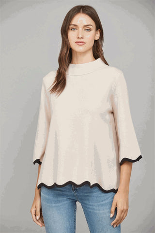 A woman wearing a blush sweater with a mock neck and scallop edge detail.