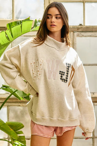 Beige sweatshirt with 'WWJD' embroidered in various patterns, featuring a mock-neck and relaxed fit worn by a model.