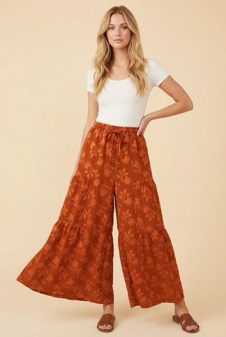 Woman wearing rust-colored wide leg lounge pants with a floral pattern, paired with a white tee and sandals.