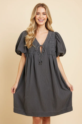 A woman wearing a relaxed-fit babydoll dress in denim with puff sleeves and a tie-front neckline.