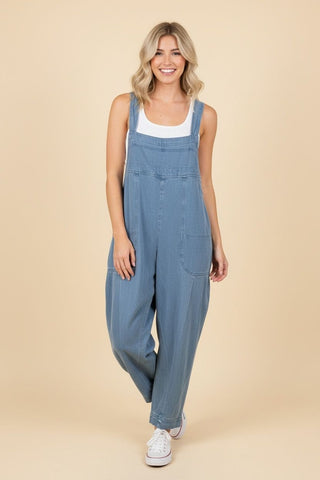 A woman wearing a relaxed fit, denim jumpsuit with shoulder straps over a white top.