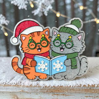 Two cartoon cats, one orange, one gray, wearing festive attire and singing from a book in a snowy setting.