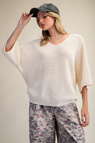 A woman wearing a natural-colored 3/4 sleeve knitted sweater from Easel, styled with a cap and patterned pants.