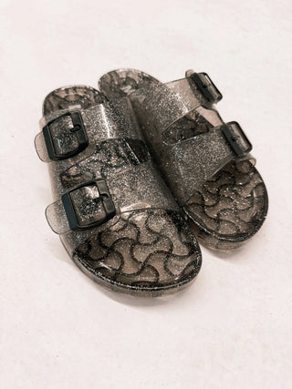 Pair of smoke-colored glitter sandals with dual adjustable buckle straps and patterned soles.