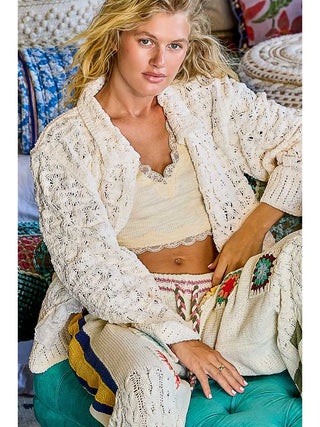 A woman wearing a cream textured crochet knit cardigan with lace trim, styled over a tank and patterned pants.