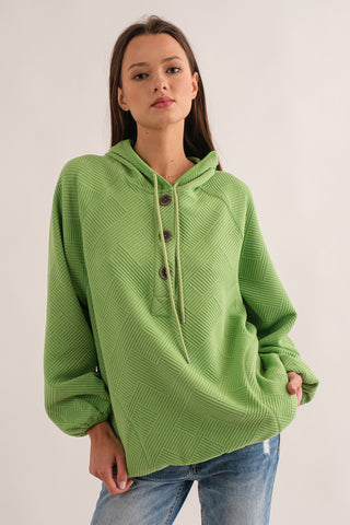 A woman wearing a green pear jacquard knit top with a hood and button detail.
