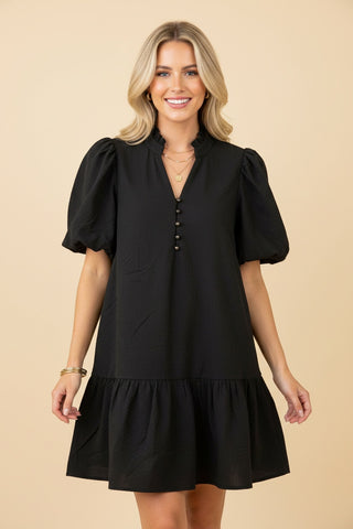 A woman wearing a black mini dress with puff sleeves and button detail.