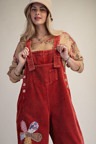 Boho red corduroy overalls with flower patch detail, worn by a person with a beige hat.
