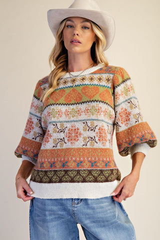 A woman wearing a multi-color knitted sweater with geometric and floral patterns in ivory, orange, and green tones.