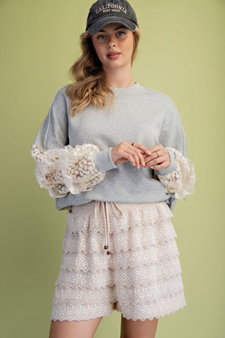 A woman wearing a heather grey pullover with lace sleeves, paired with lace shorts and a cap.