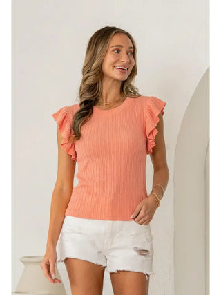Woman wearing a coral ruffled top and white shorts against a white wall.