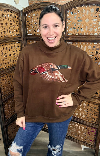A brown sweatshirt with an embroidered image of a mallard duck, worn by a person.