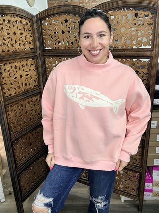 A woman wearing a pink sweatshirt with a fish graphic design, featuring a relaxed fit and mock neck.