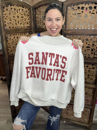 A woman holding a reversible mockneck sweater with 'Santa's Favorite' text in red on a light background.