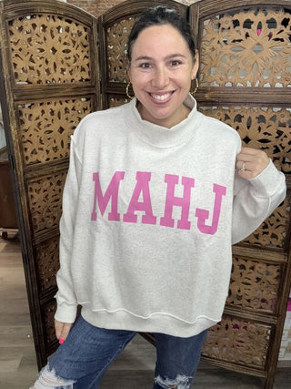 A woman wearing a light gray mockneck sweatshirt with 'Mahj' in pink text.
